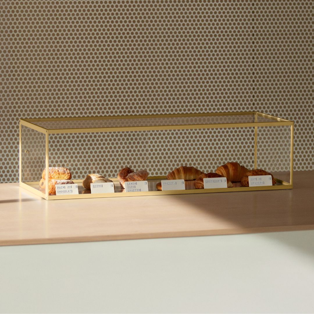 acrylic display case with pastries on a wooden shelf against a patterned wall.