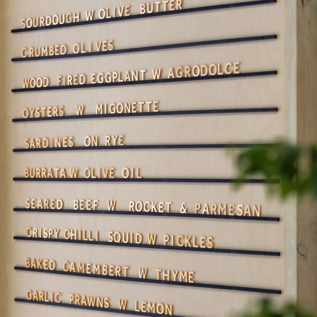 Metal menu with black rails and brass letters, mounted on wall with café menu beside green plant