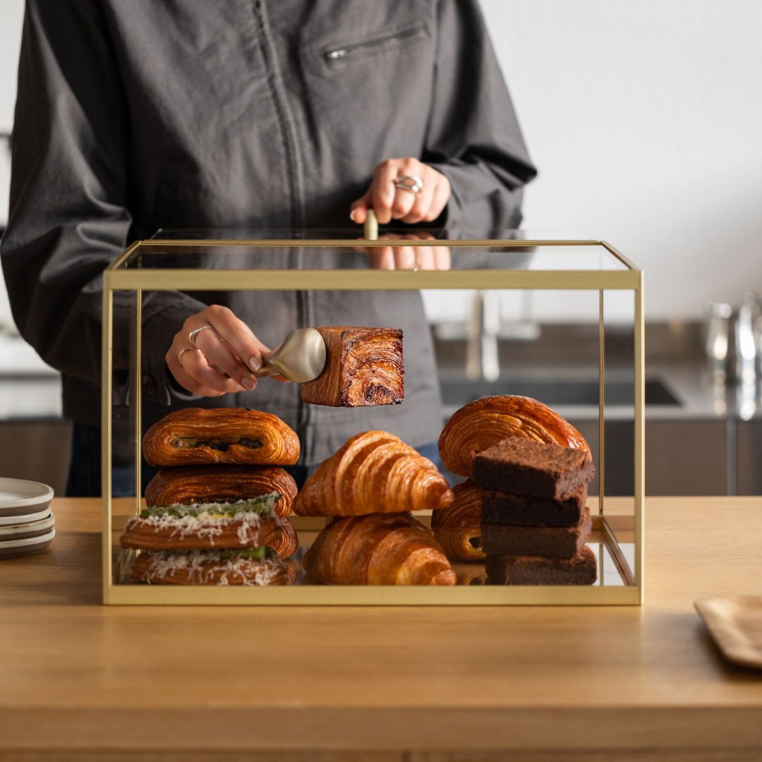 Close-up view of bakery display case showing croissants behind clear acrylic panel with person removing a pastry