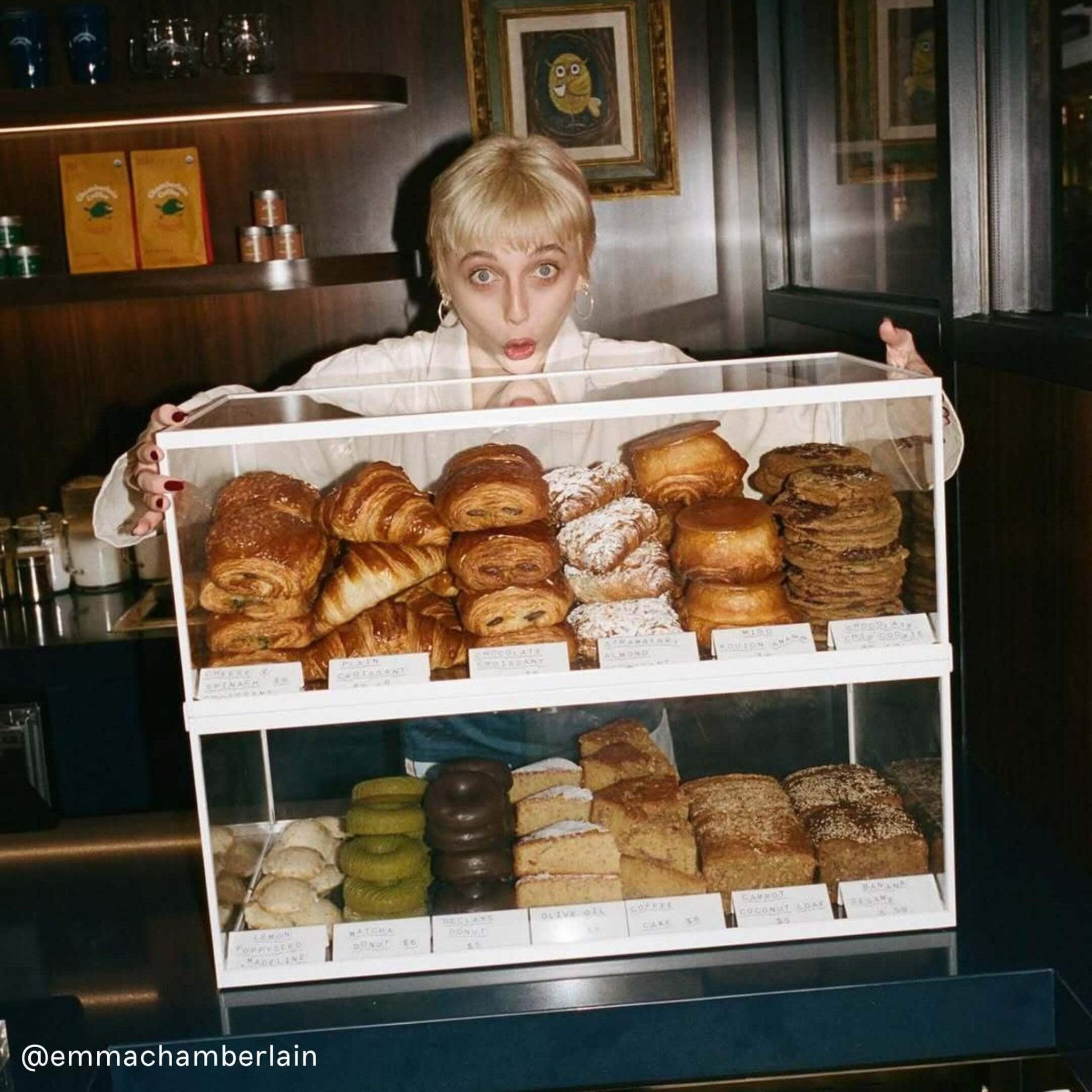 emma chamberlain standing behind a two tier bakery display case stacked with pastries and baked goods