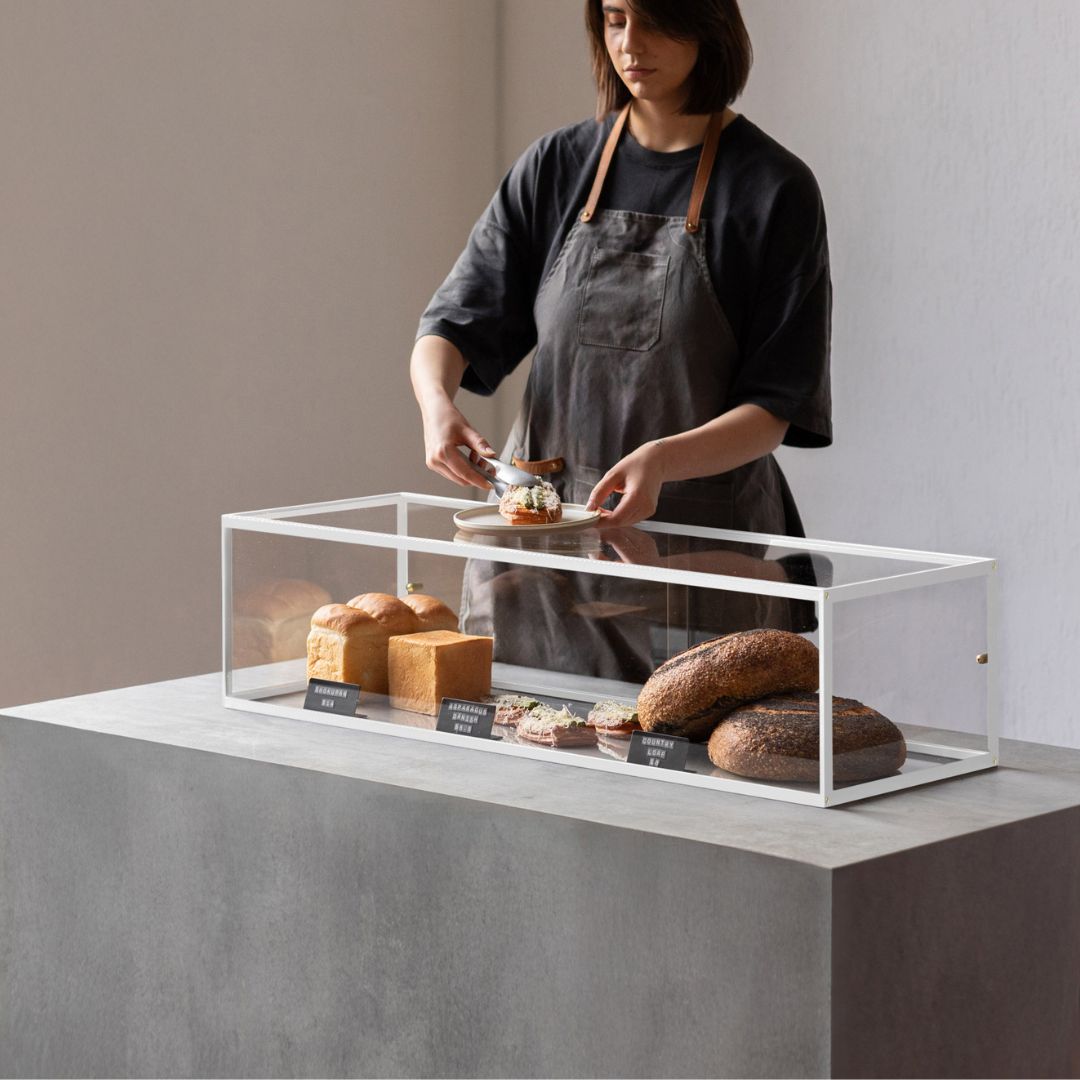 white bakery display case with breads inside and small black signs and a woman putting bread on a plate.