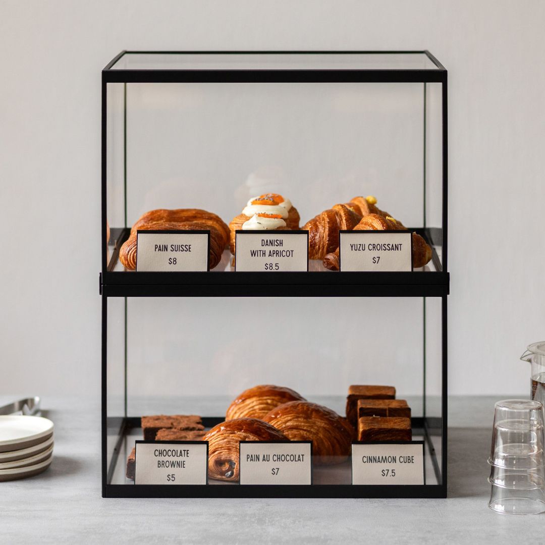black card counter signs in two stacked bakery display cases showing pastry pricing