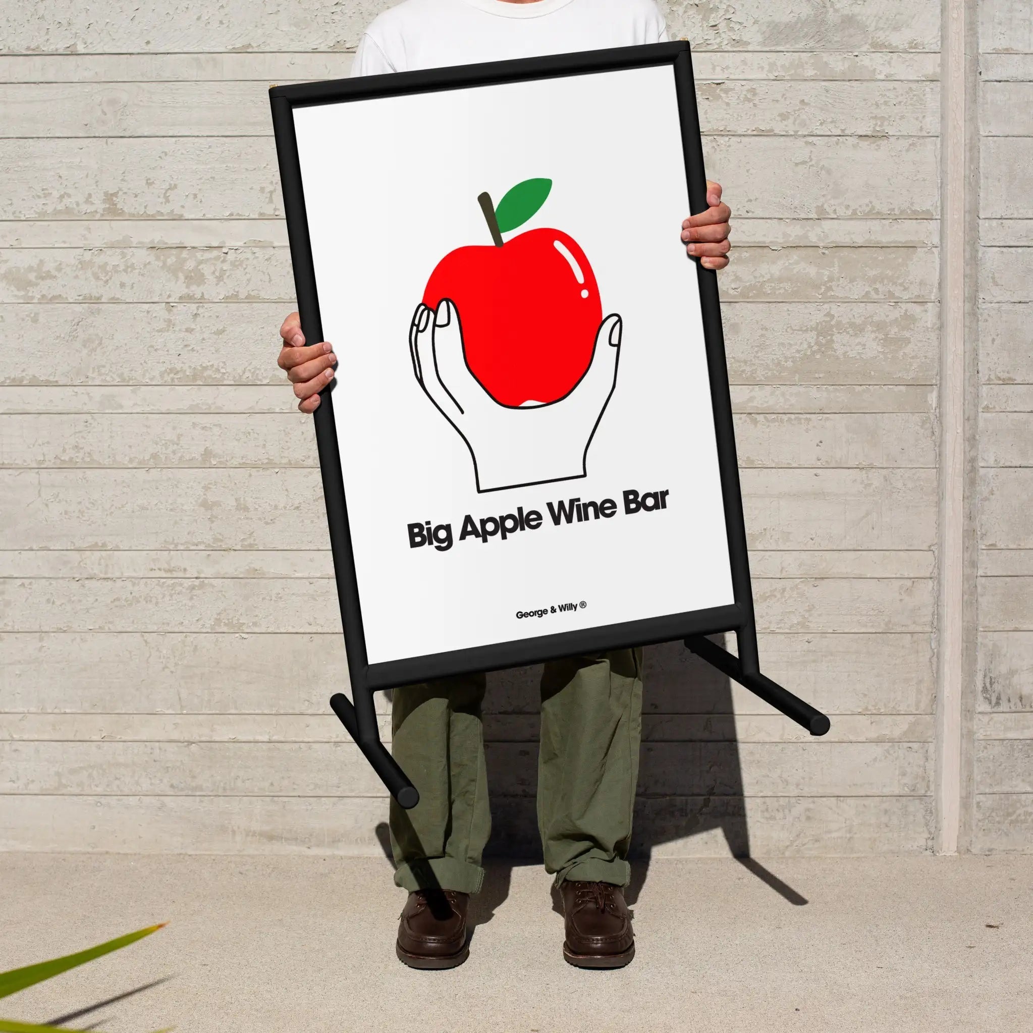 person standing on pavement holding up black framed poster sidewalk sandwich board sign with a poster of an apple inside