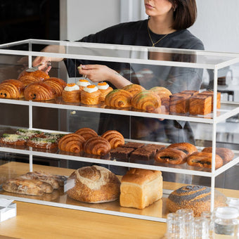 white three-tier acrylic display cabinet with pastries inside on a countertop with a staff member reaching inside