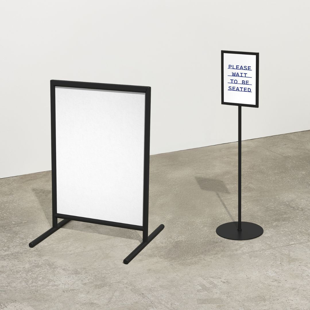 Two black poster stands with white screens on a concrete floor against a white wall.