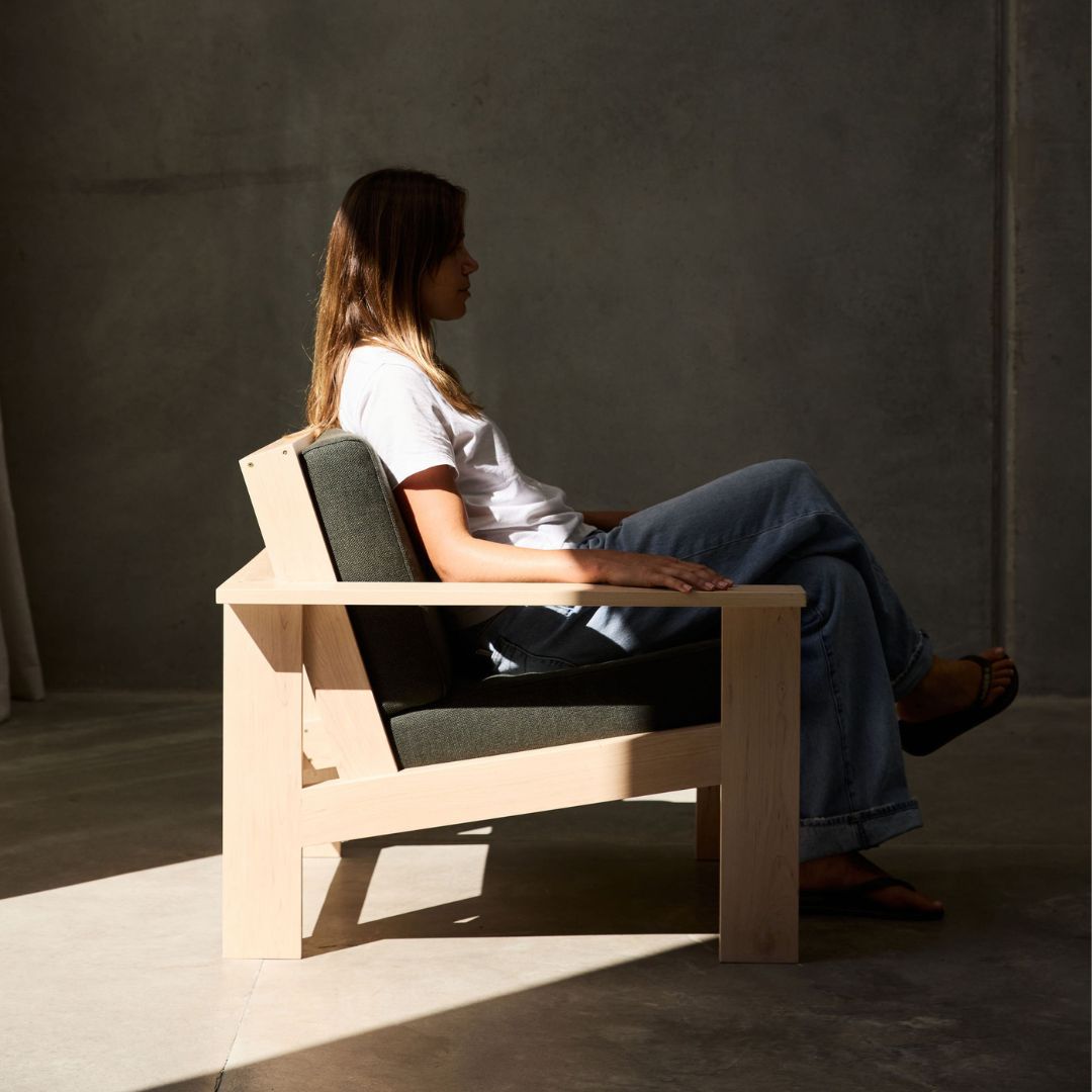 Person sitting on a modern wooden chair with a gray cushion squab in a dimly lit room.